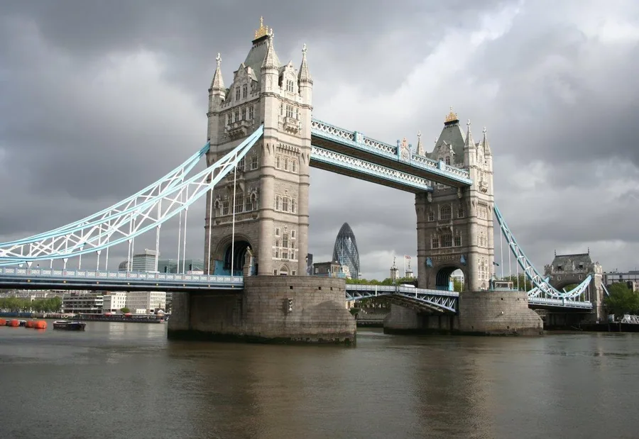 Tower Bridge London spanning the River Thames with ornate stone towers and light blue steel suspension cables against cloudy sky