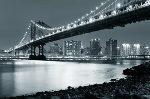Night cityscape with illuminated bridge, skyscrapers, and calm water in black and white tones.