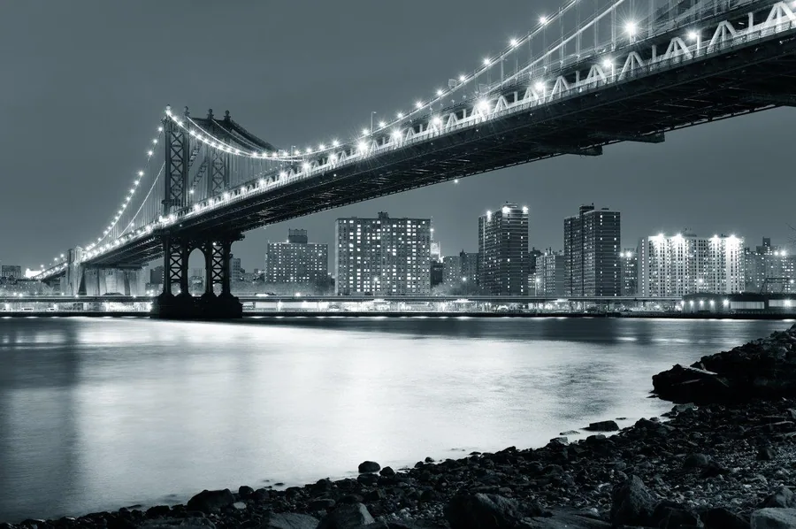 Night cityscape with illuminated bridge, skyscrapers, and calm water in black and white tones.