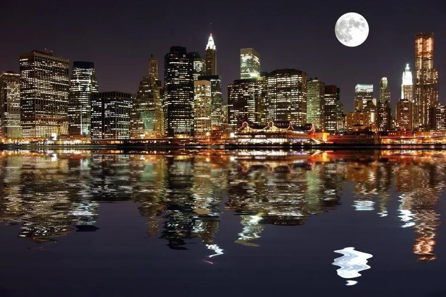 New York City skyline at night with glowing buildings reflected in water and full moon above