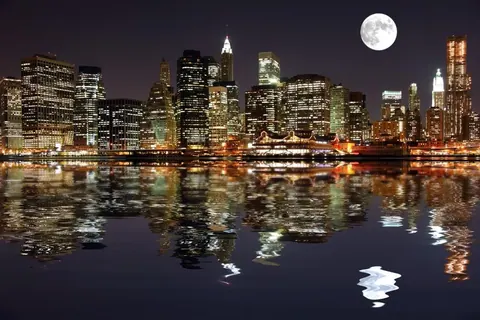 New York City skyline at night with glowing buildings, moon, and water reflections
