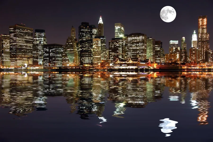 New York City skyline at night with glowing buildings, moon, and water reflections