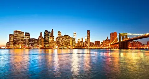 Illuminated New York City skyline at dusk with Brooklyn Bridge reflected in calm blue water under evening sky