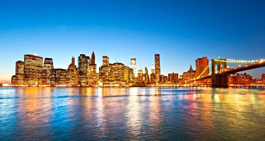 Illuminated New York City skyline at dusk with Brooklyn Bridge reflected in calm blue water under evening sky