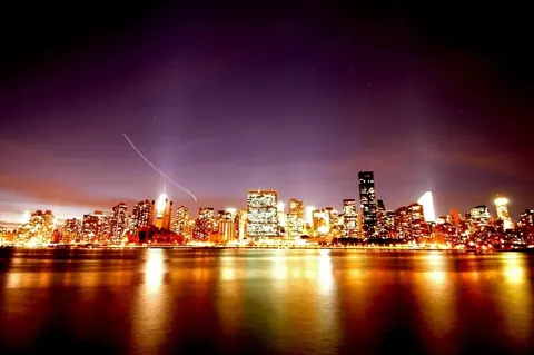 Illuminated city skyline at dusk reflected in water with crescent moon and purple sky