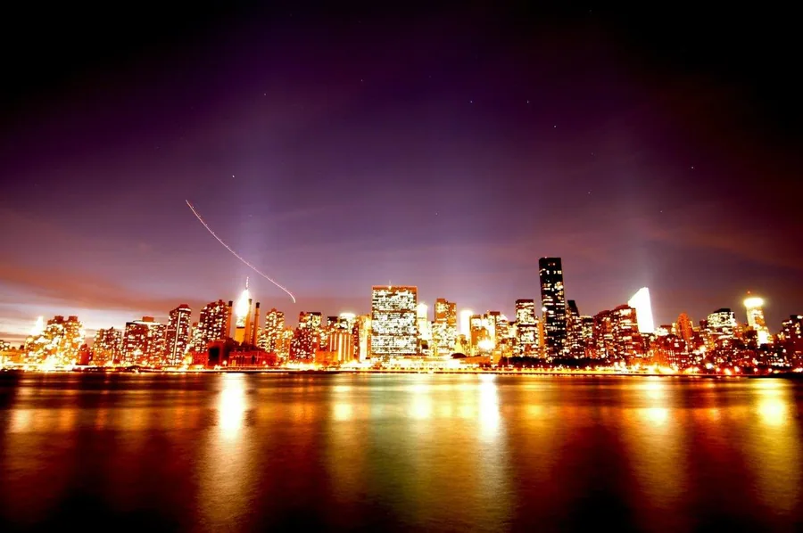 Illuminated city skyline at dusk reflected in water with crescent moon and purple sky