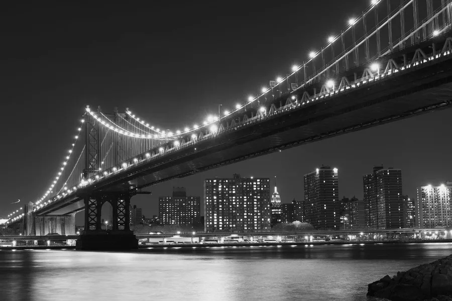 Black and white night cityscape with lit suspension bridge and illuminated high-rise buildings reflected in water.