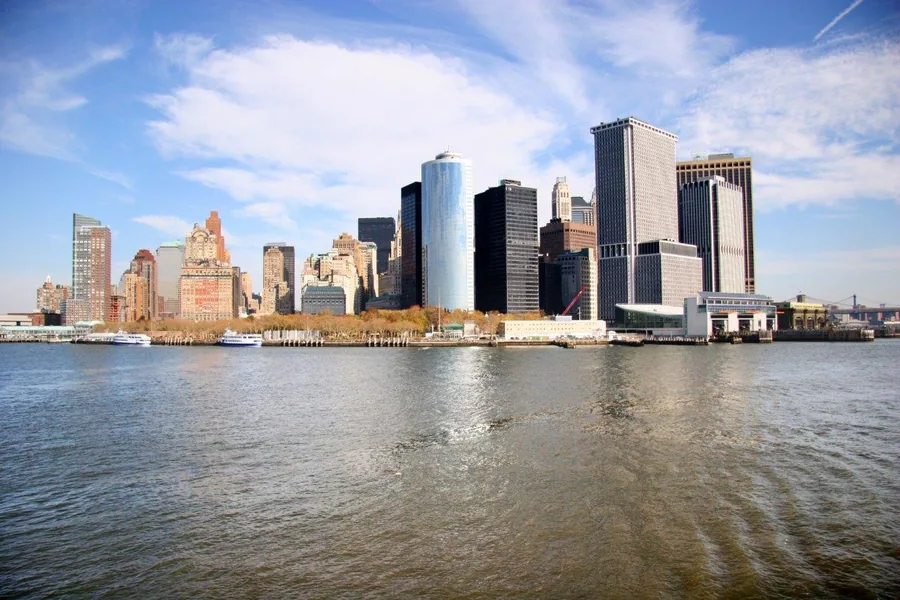 New York City skyline with tall buildings reflecting in water under blue sky with clouds