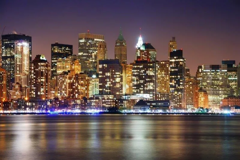 Illuminated Manhattan skyline at dusk with buildings reflected in water under purple sky
