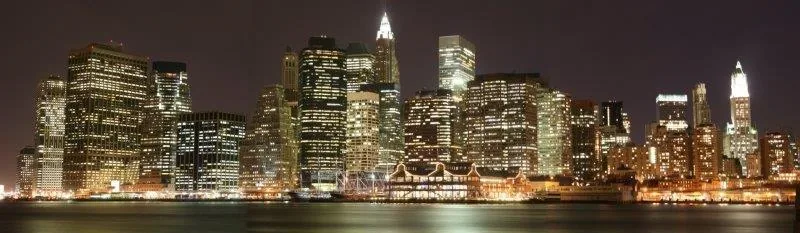 Nighttime Manhattan skyline with glowing buildings and water reflections against dark sky.