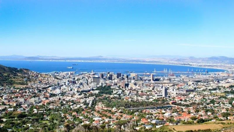 Aerial view of coastal city with blue bay, downtown skyline, and residential neighborhoods stretching to distant hills.