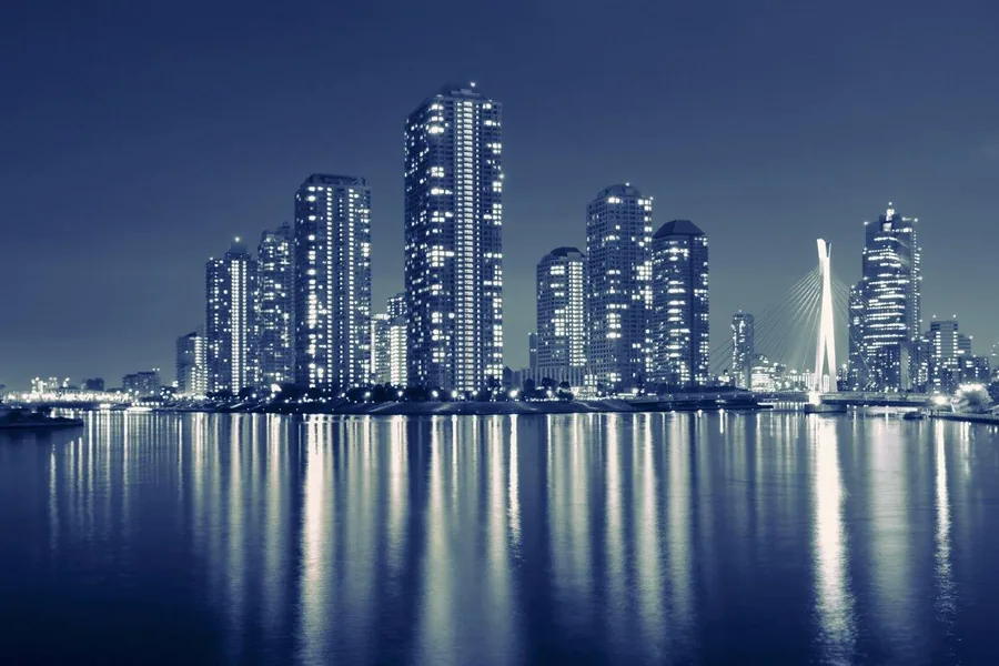 Glowing city skyline at night with tall buildings reflected in still blue water under clear sky.
