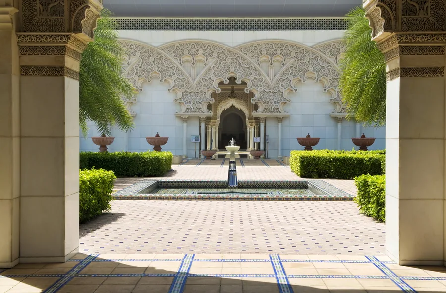 Symmetrical Moroccan courtyard with ornate archway, blue and white zellige tilework, emerald plants, and central fountain.