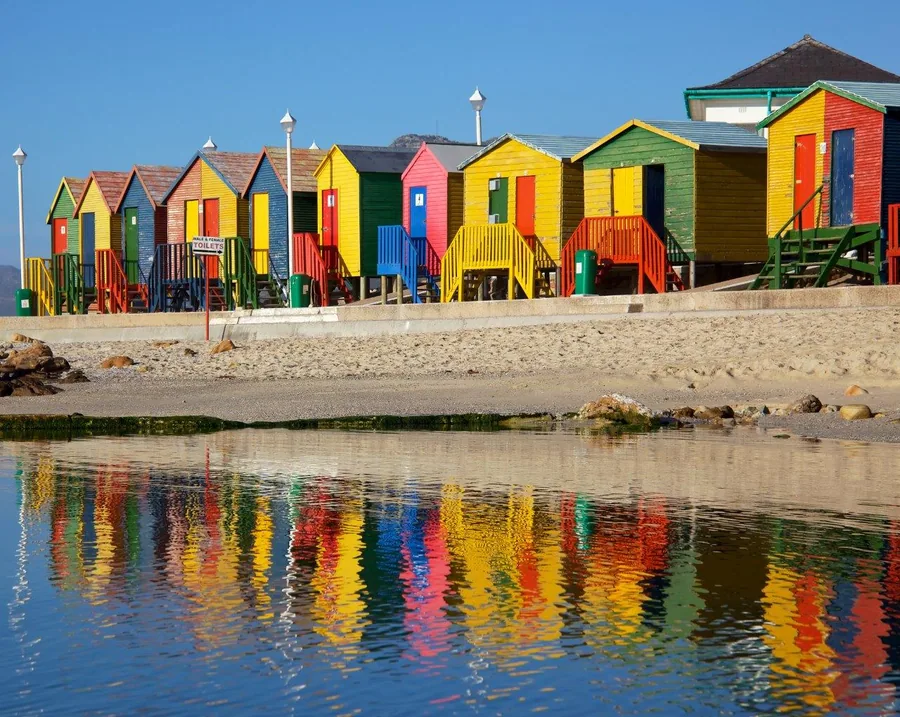 Colorful beach huts in primary colors reflected in still water, clear blue sky backdrop.
