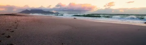 Tranquil beach at sunset with waves and mountains silhouetted against peachy-orange sky