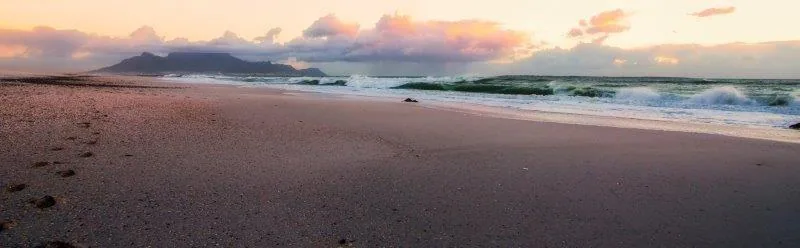 Tranquil beach at sunset with waves and mountains silhouetted against peachy-orange sky