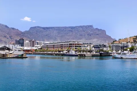 Cape Town waterfront with Table Mountain backdrop, modern buildings, boats, and blue harbor water.