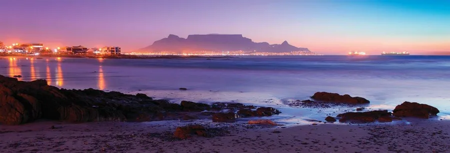 Table Mountain at dusk with purple twilight sky, city lights, and rocky coastline reflected in calm ocean waters.