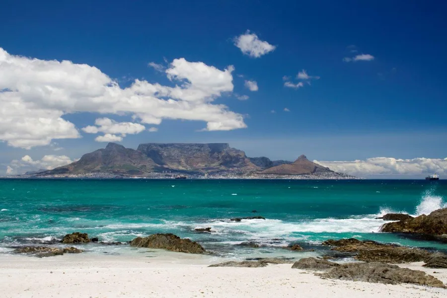 Coastal beach view with turquoise ocean, white sand, rocky shores, and a distinctive flat-topped mountain under blue sky with white clouds.