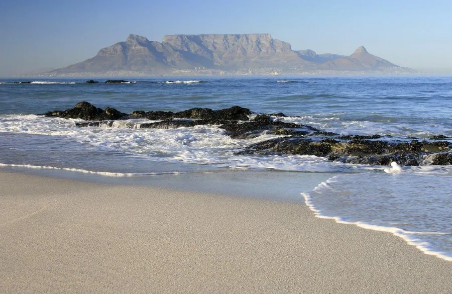 Scenic beach view with Table Mountain in the distance, white sand, turquoise waves, and dark rocks.