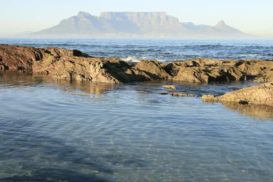 Rocky beach with calm blue ocean and flat-topped mountain in the distance under clear sky