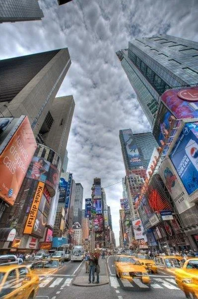 Upward view of Times Square with tall buildings, colorful advertisements, yellow cabs, and dramatic cloudy sky
