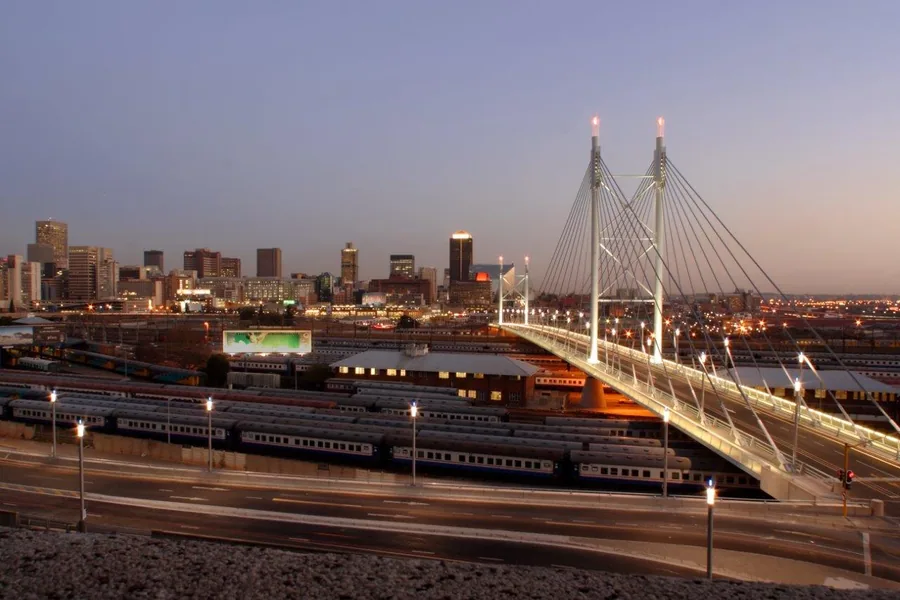 Illuminated suspension bridge with city skyline at twilight, trains and urban architecture below.