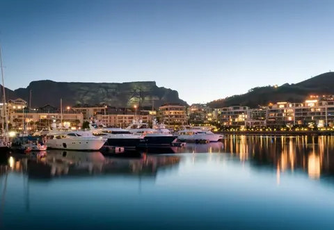 Evening harbor scene with illuminated boats, mountain backdrop, and city lights reflecting on calm water