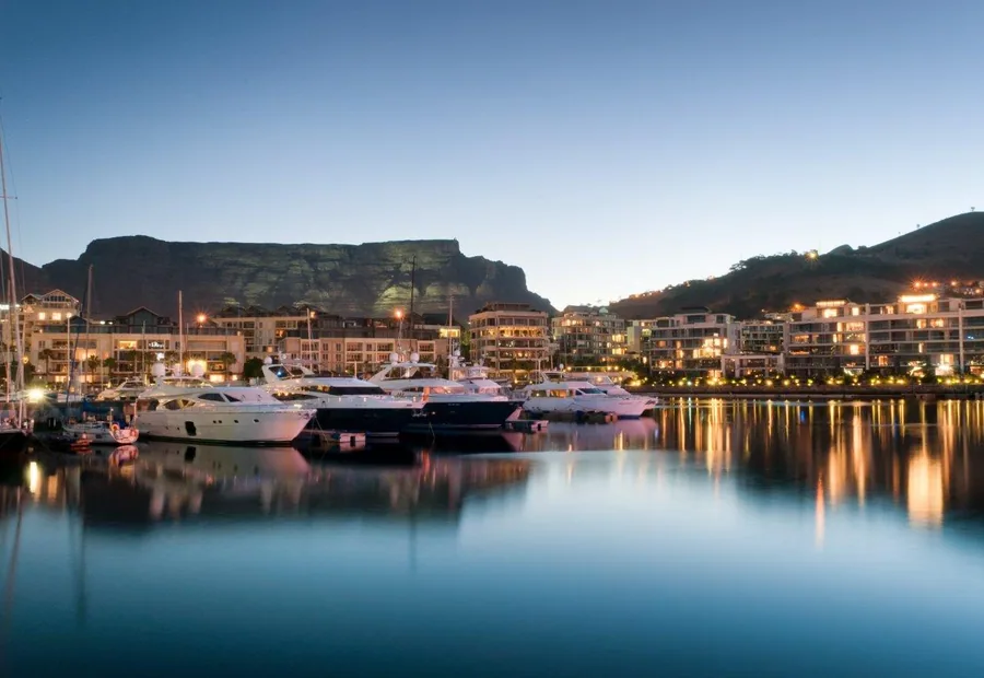 Evening harbor scene with illuminated boats, mountain backdrop, and city lights reflecting on calm water