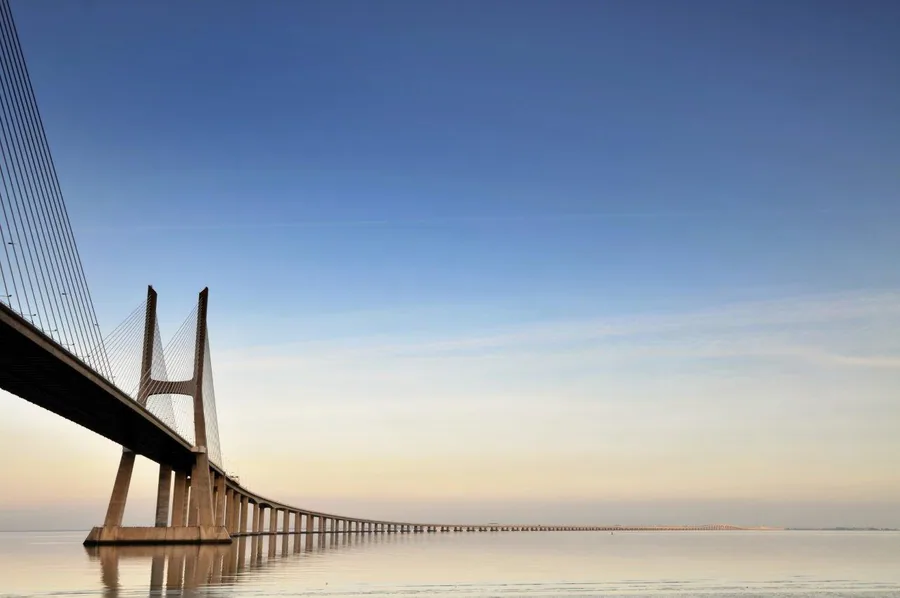 Long cable-stayed bridge extending across still water at sunset with clear sky