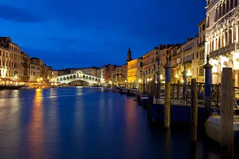 Nighttime Venice Grand Canal with Rialto Bridge, lit buildings, and mooring poles reflected in blue water at dusk