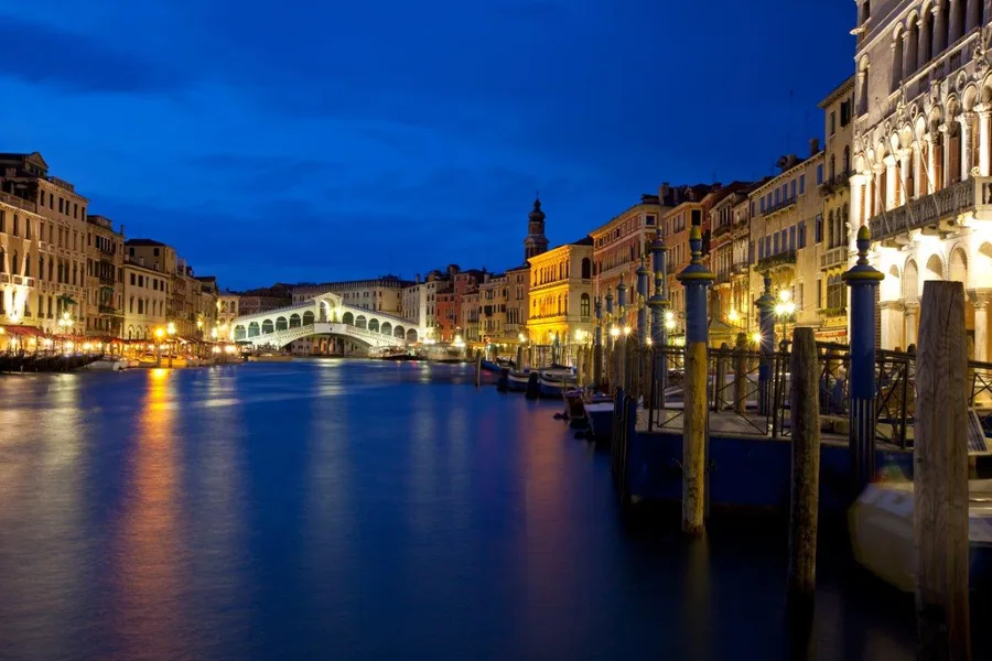 Nighttime Venice Grand Canal with Rialto Bridge, lit buildings, and mooring poles reflected in blue water at dusk