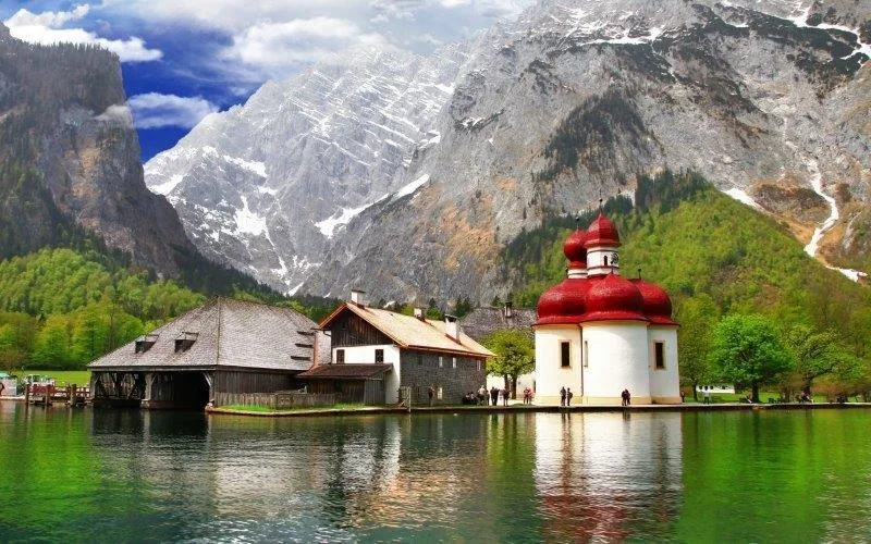 Alpine lake with white chapel with red roof, wooden boathouse, snow-capped mountains, and green forest valley