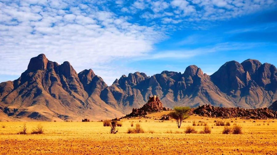 Golden desert plain with dramatic striped mountain range under blue sky with white clouds