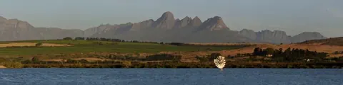 Mountain landscape with dramatic peaks reflected in river, green fields and trees in foreground.