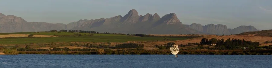 Mountain landscape with dramatic peaks reflected in river, green fields and trees in foreground.