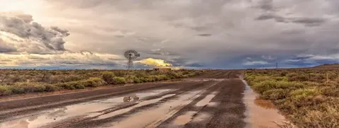 Wet dirt road stretching through semi-arid bushveld with windmill and dramatic storm clouds at sunset