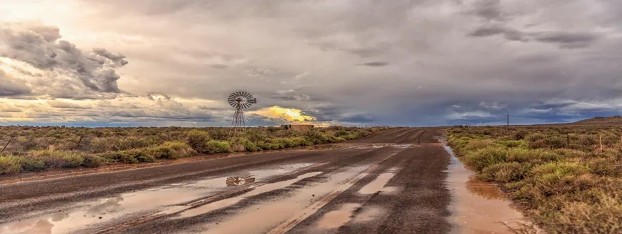 Wet dirt road stretching through semi-arid bushveld with windmill and dramatic storm clouds at sunset