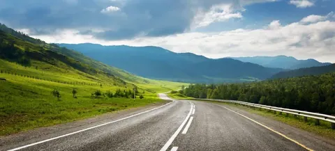 Straight asphalt road stretching through verdant valley with forested mountains and cloudy sky