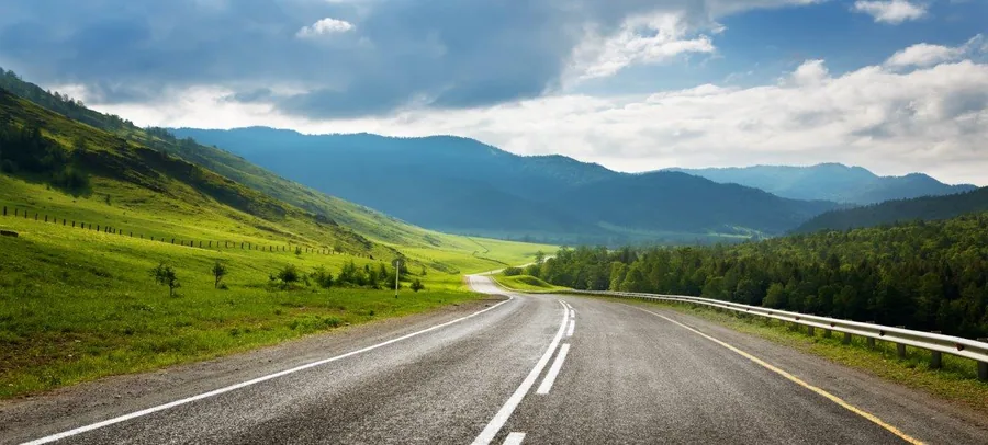 Straight asphalt road stretching through verdant valley with forested mountains and cloudy sky
