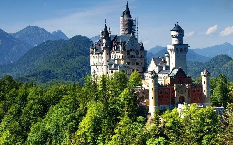 Grand castle with white and red stone towers surrounded by dense green forests and snow-capped mountains under blue sky.