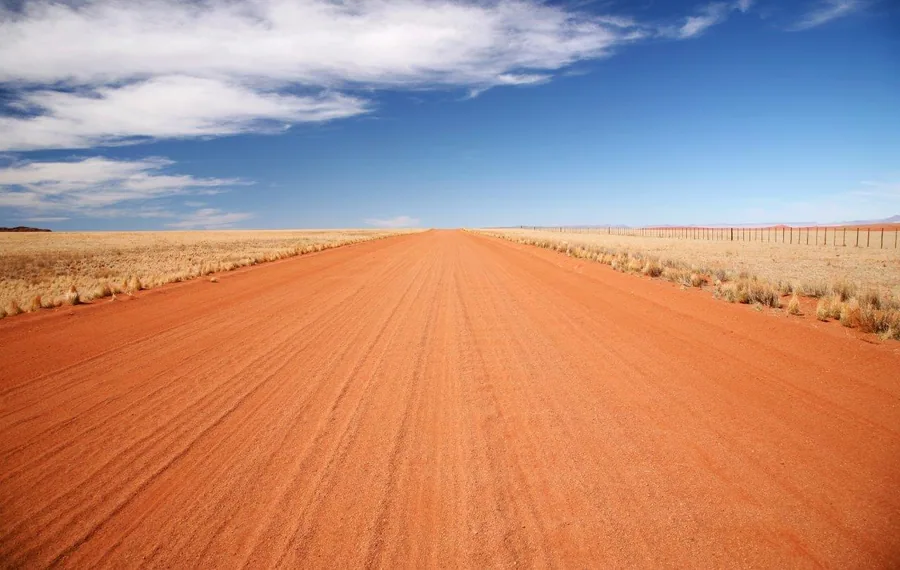 Long straight red dirt road through sparse grassland with blue sky and clouds