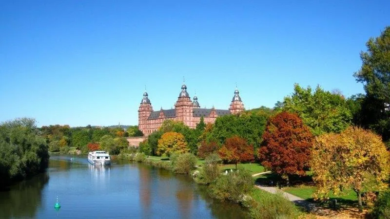 Red brick castle with spires reflected in river, surrounded by golden and crimson autumn trees under clear blue sky.