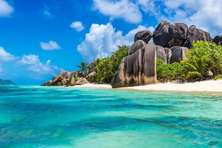 Tropical beach with massive granite rock formations, white sand, turquoise ocean, and palm trees under blue sky.