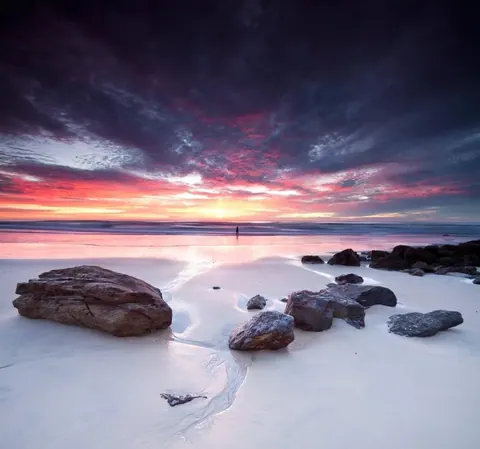 Dramatic sunset beach scene with pink and orange sky, wet sand, and scattered rocks along the shoreline.