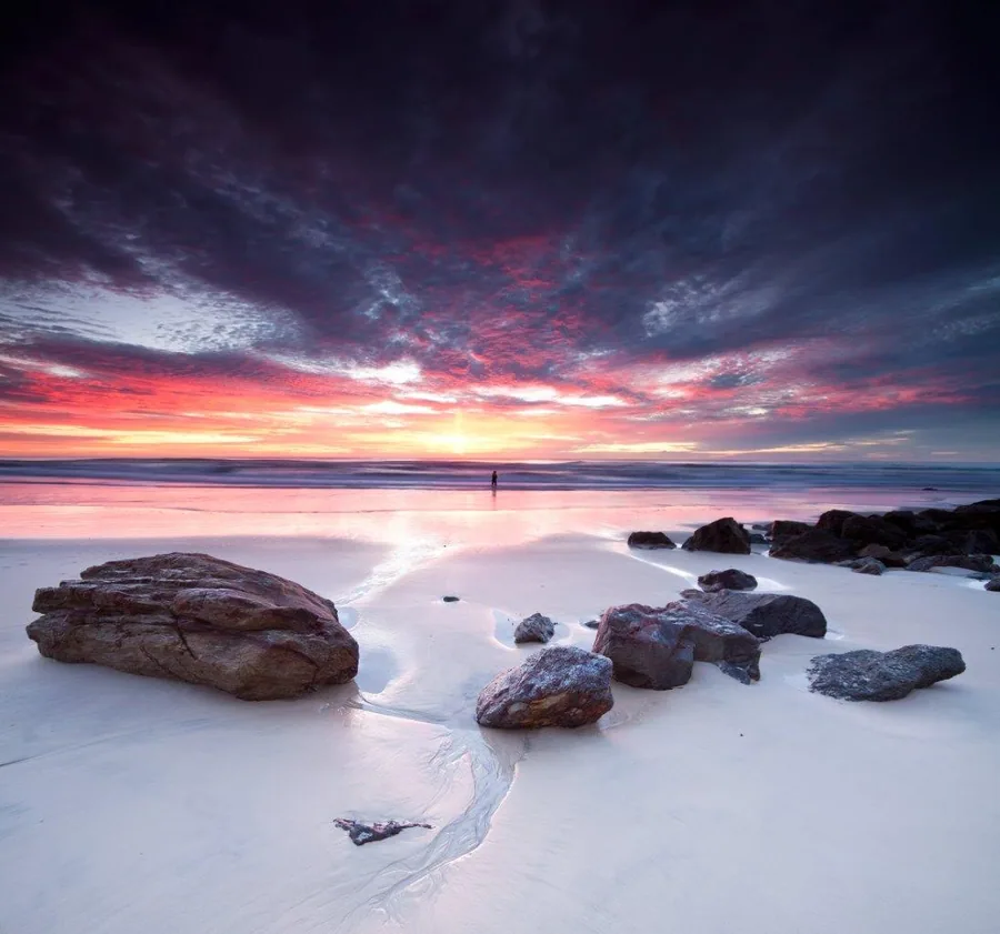 Dramatic sunset beach scene with pink and orange sky, wet sand, and scattered rocks along the shoreline.