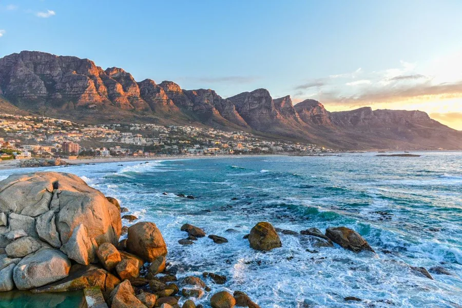 Sunset view of Table Mountain and coastal town with rocky foreshore and turquoise ocean waves.