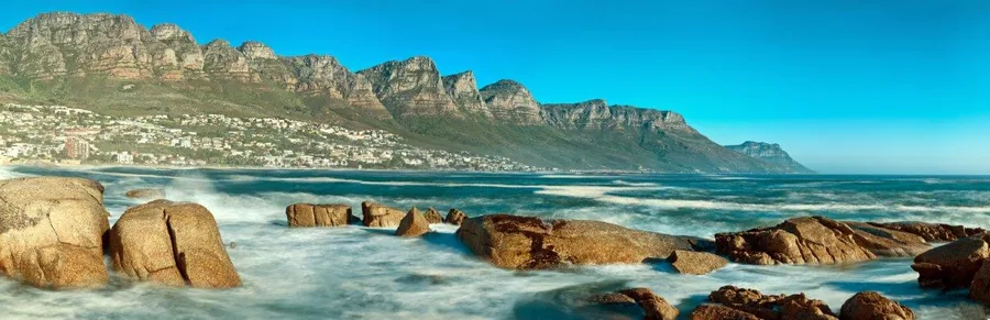 Scenic coastal beach with large granite rocks, turquoise ocean, and dramatic mountain backdrop under blue sky.