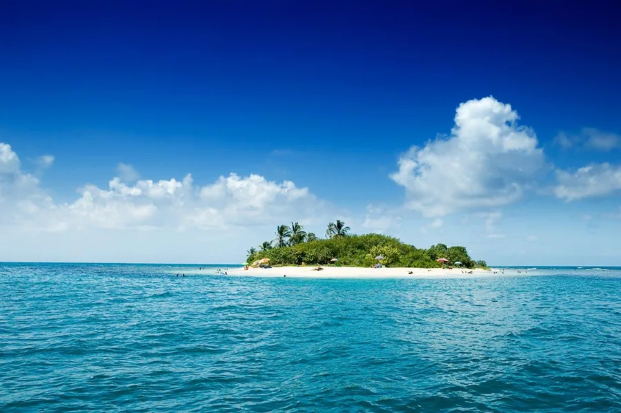 Sandy tropical island with palm trees surrounded by turquoise ocean water and blue sky with white clouds