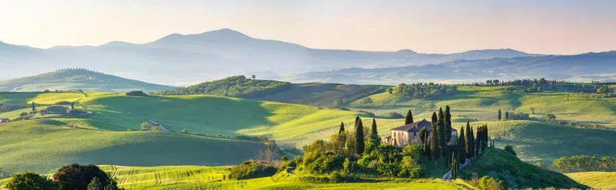 Panoramic Tuscan hillside landscape with cypress trees, villa, green fields, and distant mountains at sunrise.